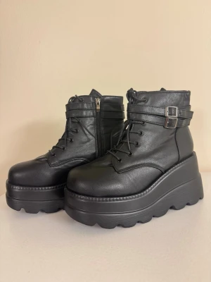 Svarta platåstövletter med spännen - Snygga svarta lätta stövletter med kraftig platåsula, snörning framtill och dubbla spännen runt ankeln. Tillverkade i konstläder med dragkedja på sidan för enkel på- och avtagning. Ger en edgy look och passar perfekt till streetwear eller rockiga outfits. Ett felköp så bara provade inomhus. Se foton för höjd på sulan.