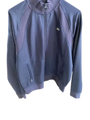 Mörkblå zip-hoodie från Lacoste - Mörkblå hoodie från Lacoste med dragkedja framtill och två fickor med dragkedja. Klassisk krokodil-logga på bröstet och hög krage med ringdetalj vid dragkedjan. Tillverkad i mjukt bomullsmaterial, perfekt för en avslappnad stil.