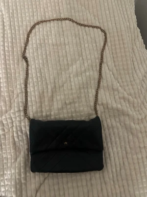 Svart axelväska med kedjerem - Snygg svart axelväska med quiltad design och guldfärgad kedjerem. Väskan har en liten guldfärgad detalj framtill och stängs med lock. Perfekt för att bära det viktigaste och ger en stilren touch till din outfit.