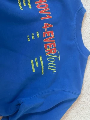 Blå Hov1 4-Ever Tour hoodie - Säljer en blå hoodie från Hov1 med trycket '4-Ever Tour' och turnédatum på ryggen samt en liten krona broderad på bröstet. Hoodien har en avslappnad passform och är gjord i mjukt bomullsmaterial. Perfekt för dig som gillar svensk hiphop och vill ha något unikt.