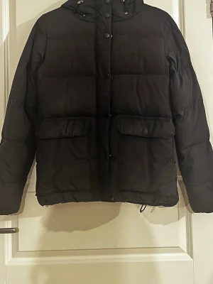 Svart dunjacka från GANT med huva - Snygg svart dunjacka från GANT med huva och stora framfickor. Jackan har dragkedja och knappar framtill samt justerbar nederkant med dragsko. Perfekt för kalla vinterdagar och riktigt skön att bära. Klassisk design som passar till det mesta.