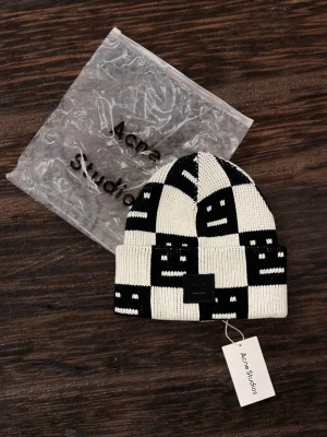 Svartvit mössa från Acne Studios - Säljer en svartvit stickad mössa från Acne Studios med rutigt mönster och deras ikoniska Face-logga framtill. Mössan har uppvikt kant och är tillverkad i mjukt material som håller dig varm och snygg. Perfekt accessoar för att lyfta din outfit.
