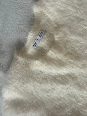 Krämvit fluffig stickad topp Zara - Supersöt krämvit stickad topp från Zara med korta ärmar och rund hals. Materialet är fluffigt och mjukt, vilket ger en mysig  look. Perfekt att styla för en cool vardagsoutfit.