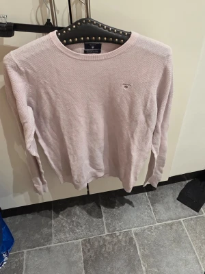 Ljusrosa stickad tröja från Gant - Ljusrosa stickad tröja från Gant med rund halsringning och diskret broderad logotyp på bröstet. Tröjan har lång ärm och är tillverkad i ett mjukt, strukturerat material som passar perfekt för lager-på-lager.