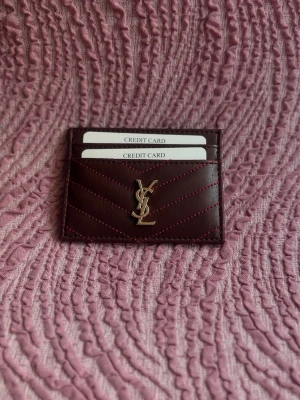 Vinröd YSL kortfodral i läder - Snyggt vinrött kortfodral från YSL i äkta läder med guldfärgad logga framtill. Fodralet har flera kortfack och dekorativa sömmar i matchande färg. Perfekt för att hålla ordning på dina kort med stil.