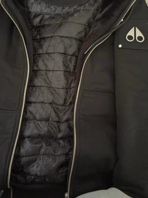 Svart dunjacka från Moose Knuckles - Snygg svart dunjacka från Moose Knuckles med quiltat innerfoder och silverfärgade dragkedjor. Jackan har en ikonisk logga på bröstet och stilren design som passar perfekt till kalla vinterdagar. Materialet är slitstarkt och håller värmen riktigt bra.