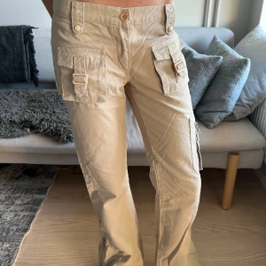 Vintage cargo pants - Snygga beige cargobyxor med flera stora fickor framtill och på sidan av benen. Byxorna har dekorativa spännen på ficklocken och vida ben. Perfekta för en avslappnad och trendig stil.