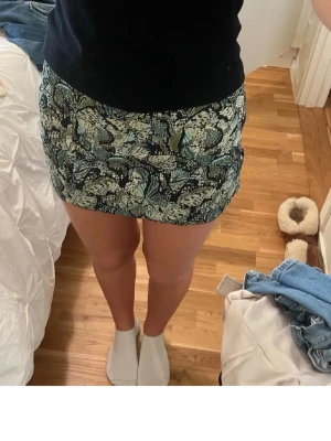 Blommig kortkjol i grönt och svart - Säljer en snygg kortkjol med blommigt mönster i gröna, vita och svarta toner. Kjolen har en tight passform och är perfekt för dig som gillar trendiga och färgglada plagg. Materialet känns mjukt och stretchigt, vilket gör den bekväm att bära.