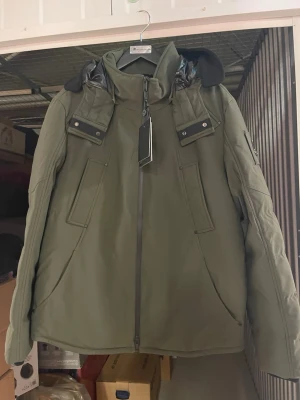 Olivgrön parkas från Moose Knuckles - Snygg olivgrön parkas från Moose Knuckles med svarta detaljer och dragkedja framtill. Jackan har flera fickor, hög krage och en huva med svarta detaljer. Metallogo på ärmen ger en exklusiv känsla. Perfekt för kalla dagar.