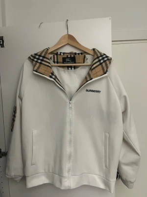 Vit ziptröja från Burberry - Vit ziptröja från Burberry med klassiskt rutigt foder i kragen och logga broderad på bröstet. Tröjan har två fickor framtill och en avslappnad passform. Perfekt för en stilren och trendig look.