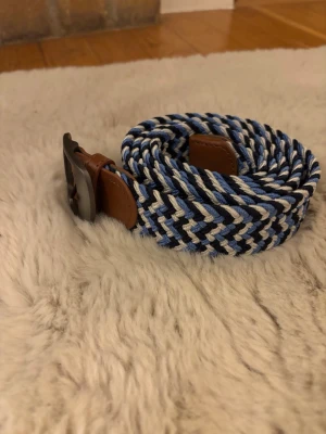 Randigt flätat bälte i blått och vitt - Snyggt flätat bälte med randigt mönster i blått, vitt och svart. Spänne och detaljer i brunt skinn ger en schysst kontrast. Perfekt accessoar för att lyfta din outfit med lite färg och stil.