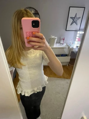 Vit topp med volangkant  - Söt vit topp med smockad framsida och volangkant nedtill. T-shirten har rund halsringning och korta ärmar. Från ginatricot