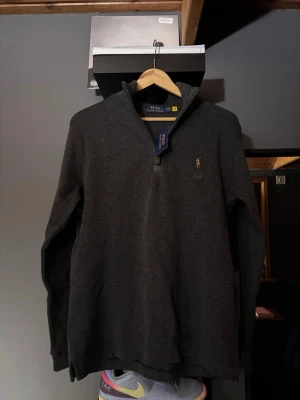 Mörkgrå halvzip från Polo Ralph Lauren - Mörkgrå långärmad tröja från Polo Ralph Lauren med halv dragkedja och klassisk broderad logga på bröstet. Tröjan har hög krage och är tillverkad i mjukt material, perfekt för lager-på-lager-stil.