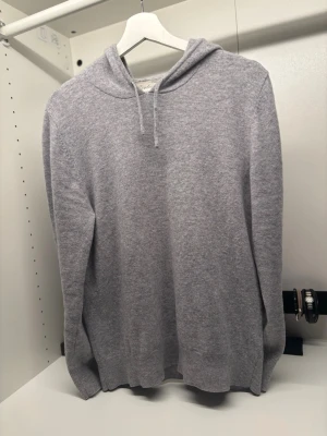 Ljusgrå stickad hoodie - Mysig ljusgrå stickad hoodie med huva och dragsnören. Tröjan har ribbade muddar vid ärmslut och nederkant, vilket ger en skön passform. Perfekt för dig som gillar en enkel och stilren look med lite extra komfort.