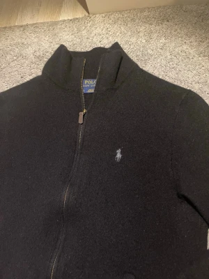 Svart ziptröja från Polo Ralph Lauren - Klassisk svart ziptröja från Polo Ralph Lauren med hög krage och dragkedja framtill. Tröjan har det ikoniska broderade logotypen på bröstet och är tillverkad i ett mjukt, stickat material. Perfekt för en stilren och avslappnad look. Tröjan är i storlek L men sitter mer som en S/M.