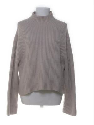 Beige ribbstickad croppad tröja - Säljer en beige ribbstickad tröja med croppad passform och långa ärmar. Tröjan har en hög hals och är tillverkad i ett mjukt, tjockare material som ger en avslappnad look. Perfekt för lager-på-lager-stil.