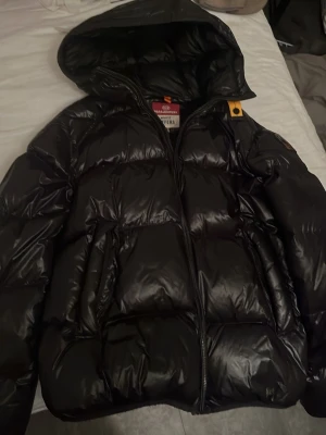 Svart pufferjacka Parajumpers - Säljer en svart glansig pufferjacka från Parajumpers, modell Buzz Puffers. Jackan har huva, dragkedja framtill och två stora fickor. Perfekt för kalla vinterdagar och riktigt snygg streetstil. Har kvitto (råka spreja lite sprejfärg på det) Passar om man är mellan 175-180cm