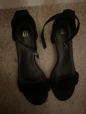 Svarta pumps med blockklack och rem - Snygga svarta pumps med bred blockklack och öppen tå. Skorna har en justerbar rem runt ankeln med silverfärgat spänne. Materialet ser ut att vara mockaimitation och de har en stilren, klassisk design som passar till många outfits.