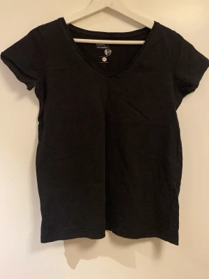 Svart V-ringad T-shirt H&M Basic - En svart, enkel T-shirt från H&M Basic med V-ringning och korta ärmar. Modellen är rak och i mjukt bomullsmaterial, perfekt för en clean och stilren look.