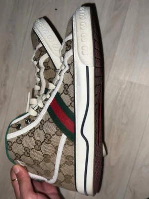 Gucci high top sneakers beige/grön/röd - Snygga high top sneakers från Gucci med klassiskt beige monogrammönster, gröna och röda ränder på sidan samt vita detaljer. Skorna har vita snören, grön insida och vit sula med GG-logga. Tillverkade i canvas och läder för en lyxig känsla.