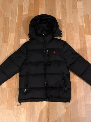 Svart dunjacka från Polo Ralph Lauren - Säljer en svart dunjacka från Polo Ralph Lauren med huva och dragkedja framtill. Jackan har två sidofickor med dragkedja och en bröstficka. Klassisk röd logga på bröstet och justerbar nederkant. Perfekt för kalla vinterdagar.