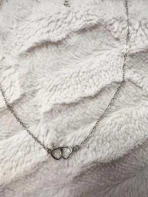 Silverhalsband med dubbla hjärtan - Sött halsband i silverfärgad metall med två sammanlänkade hjärtan som hänge. Kedjan är tunn och har ett justerbart lås. Perfekt för dig som gillar minimalistiska och romantiska smycken.