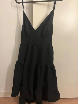 Svart klänning med volang - svart klänning med smala axelband, v-ringning och rosett i ryggen. Klänningen har volanger i kjolen. Har aldrig andvänt 