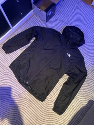 Svart regnjacka/vindjacka  från The North Face - Snygg svart regnjacka från The North Face med huva och dragkedja framtill. Jackan har en minimalistisk design med logga på bröstet och är tillverkad i lätt, vattenavvisande material. Perfekt för regniga dagar när du vill hålla dig torr och stilren. Oanvänd sprillans ny