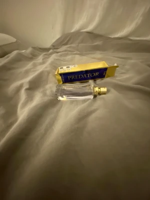 Predator parfym med guldlock - Säljer en parfym från Predator med elegant, genomskinlig glasflaska och stilrent guldigt spraylock. Förpackningen är blå och gul med tydlig logga. Perfekt för dig som vill ha en unik doft i snygg design.