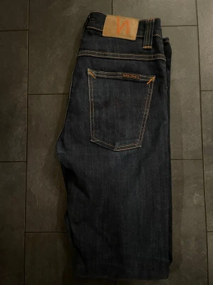 Mörkblå jeans från Nudie Jeans  - Säljer ett par mörkblå jeans från Nudie Jeans  med klassisk femficksdesign och kontrastsömmar i orange. Jeansen har rak passform och är tillverkade i slitstarkt denimtyg av bomull. Märkespatch i läder baktill och snygga detaljer på bakfickorna.kan gå ner i pris vid snabb affär 