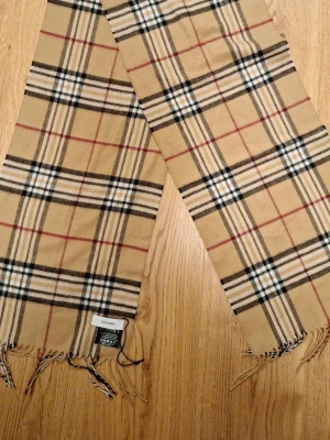 Burberry liknande halsduk - Klassisk beige halsduk från Don Donna med rutigt mönster i svart, vitt och rött. Halsduken har fransar i ändarna och är tillverkad i mjuk polacryl (coshmink). Perfekt accessoar för att lyfta din outfit med en tidlös look. Säljs flr att den inte kommer till annvändning