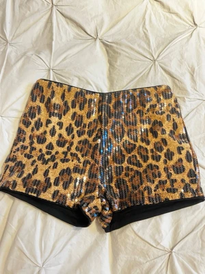 Leopardmönstrade paljettshorts - Snygga shorts med leopardmönster och glittriga paljetter. De har en hög midja och kort passform, perfekta för dig som vill sticka ut. Färgerna går i beige, brun och svart med en skimrande finish.