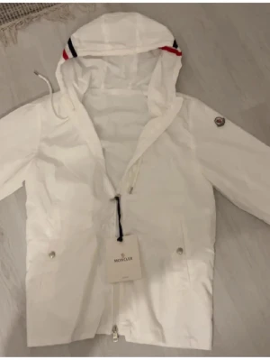 Vit vindjacka från Moncler med huva - Snygg vit vindjacka från Moncler med huva och dragkedja framtill. Jackan har Moncler-logga på ärmen, två fickor med knappar och coola rödblå detaljer på huvan. Storlek 2 alltås M men passar 100% S. 😀.