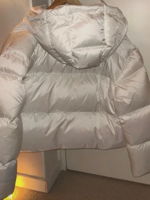 Vit pufferjacka med huva - Snygg vit pufferjacka från Busnel,  med huva och dragkedja framtill. Jackan har en quiltad design och två fickor med knappstängning. Perfekt för kyliga dagar och ger en trendig look.  Nypris 6500kr