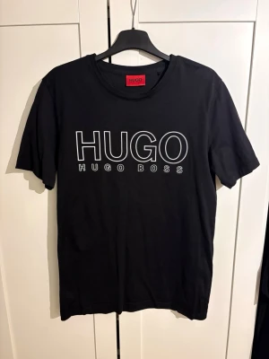 Svart HUGO BOSS t-shirt  - Svart t-shirt från HUGO BOSS med stort vitt HUGO-tryck på bröstet. Klassisk rund halsringning och korta ärmar. Perfekt för en stilren och modern look. Tillverkad i mjuk bomull för extra komfort.