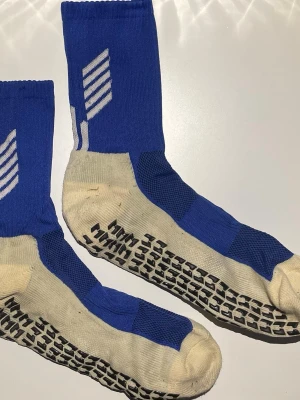 Blå-vita sportstrumpor med grepp - Säljer ett par blå och vita sportstrumpor med svart greppmönster undertill. Strumporna har en sportig design med vita detaljer och är perfekta för dig som vill ha extra grepp vid träning eller fotboll. Materialet är bomull och syntet för bästa komfort.
