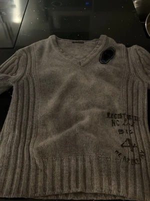 Grå stickad tröja med tryck och patch - Grå stickad tröja med V-ringning och ribbade detaljer. Tröjan har ett svart tryck nertill på framsidan och en svart patch på vänster axel. Snyggt mönster i stickningen och lång ärm. Perfekt för dig som gillar streetstyle. Riktigt grich