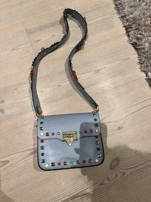 Ljusblå axelväska med nitar och blommor - Säljer en ljusblå axelväska i skinnimitation med färgglada nitar i rött, grönt och guld runt locket. Väskan har ett guldfärgat spänne och en bred axelrem dekorerad med blommor i olika färger. Köpt i Spanien och använd fåtal gången!
