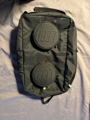 Svart ryggsäck från lego med rund design - Cool svart ryggsäck från 0937 med två stora runda detaljer framtill och vadderade axelband. Väskan har en unik form och tydliga loggor på både framsidan och axelbanden. Tillverkad i slitstark textil och har praktiska spännen och dragkedjor.