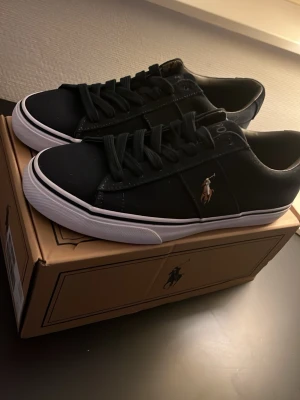 Marinblå sneakers från Polo Ralph Lauren - 🐎Stilrena svarta sneakers från Polo Ralph Lauren med klassisk låg profil och vit sula. Skorna har svart canvasmaterial, svarta skosnören och den ikoniska broderade polospelaren i brunt och vitt på sidan. Perfekta för en clean och avslappnad look.