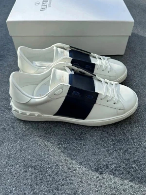 Vita sneakers från Valentino Garavani - Snygga vita sneakers från Valentino Garavani med bred mörkblå läderdetalj över snörningen och ikoniska nitar på hälen. Skorna har en clean design med vita skosnören och låg profil. Perfekta för dig som gillar exklusiv och stilren streetwear.
