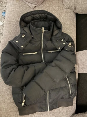 Svart dunjacka från Moose Knuckles - Snygg svart dunjacka från Moose Knuckles med avtagbar huva, silverfärgade dragkedjor och två bröstfickor. Jackan har ribbade muddar och ikoniska metallogo på ärmen. Perfekt för kalla vinterdagar och riktigt stilren look. Den är äkta och är köpt för ca 12000 kr förra vintern så den är i bra skick. Priset kan diskuteras vid snabba affärer.