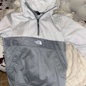 Grå och vit hoodie från The North Face - Säljer en snygg hoodie från The North Face i tvåfärgad design med ljusgrå och vit färg. Tröjan har huva, halv dragkedja och magficka. Perfekt för kyliga dagar och har en sportig vibe. Logga på bröstet och ryggen.