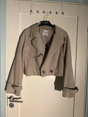 Kort beige trenchcoat från Bershka - Trendig kort trenchcoat från Bershka i beige bomullsmix. Jackan har breda slag, dubbelknäppning framtill och justerbara remmar vid ärmsluten. Perfekt för dig som vill ha en modern twist på den klassiska trenchcoaten.