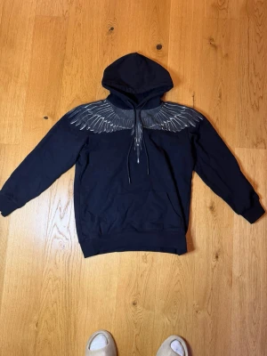 Svart hoodie Marcelo Burlon vingar - Säljer en svart hoodie från Marcelo Burlon County of Milan med ikoniskt vingtryck över axlar och bröst. Hoodien har huva med dragsko och en stor magficka. Tillverkad i mjuk bomull, perfekt för en streetwear-look.