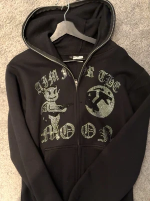 Svart hoodie Aim For The Moon, S - Svart hoodie med dragkedja från Aim For The Moon. Framsidan har blingade detaljer med texten 'Aim For The Moon' och motiv av en tecknad figur och måne. Hoodien har huva och är tillverkad i mjukt material. 