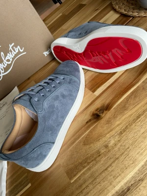 Blåa Louboutin sneakers i mocka - Snygga blåa sneakers från Christian Louboutin i mjuk mocka med klassisk röd sula och vita kanter. Skorna har lågt skaft, ton-i-ton snörning och diskret logga på sulan. Perfekta för dig som vill sticka ut med exklusiv stil. Helt nya aldrig använda 10/10 skick. Allt ingår 