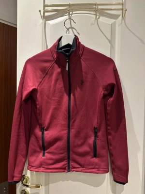 Vinröd ziptröja från Peak Performance - Snygg vinröd ziptröja från Peak Performance med svart dragkedja och två fickor framtill. Tröjan har hög krage och diskret logga på bröstet och ärmen. Perfekt för lager-på-lager och chill dagar. Materialet känns mjukt och stretchigt.