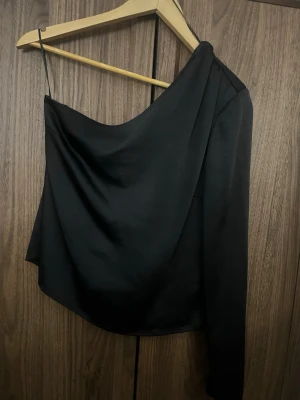 Svart oneshoulder topp Gina Tricot - Snygg svart oneshoulder topp från Gina Tricot med en lång ärm och en smal axelrem. Toppen har en draperad look och är perfekt för dig som vill ha något trendigt och stilrent. Materialet känns mjukt och följsamt.