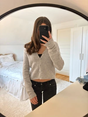 Grå stickad kofta  - Mysig grå kofta i en mjuk, stickad ullblandning. Croppad modell med rund halsringning och knäppning framtill. Perfekt att styla med jeans eller kjol för en avslappnad look.